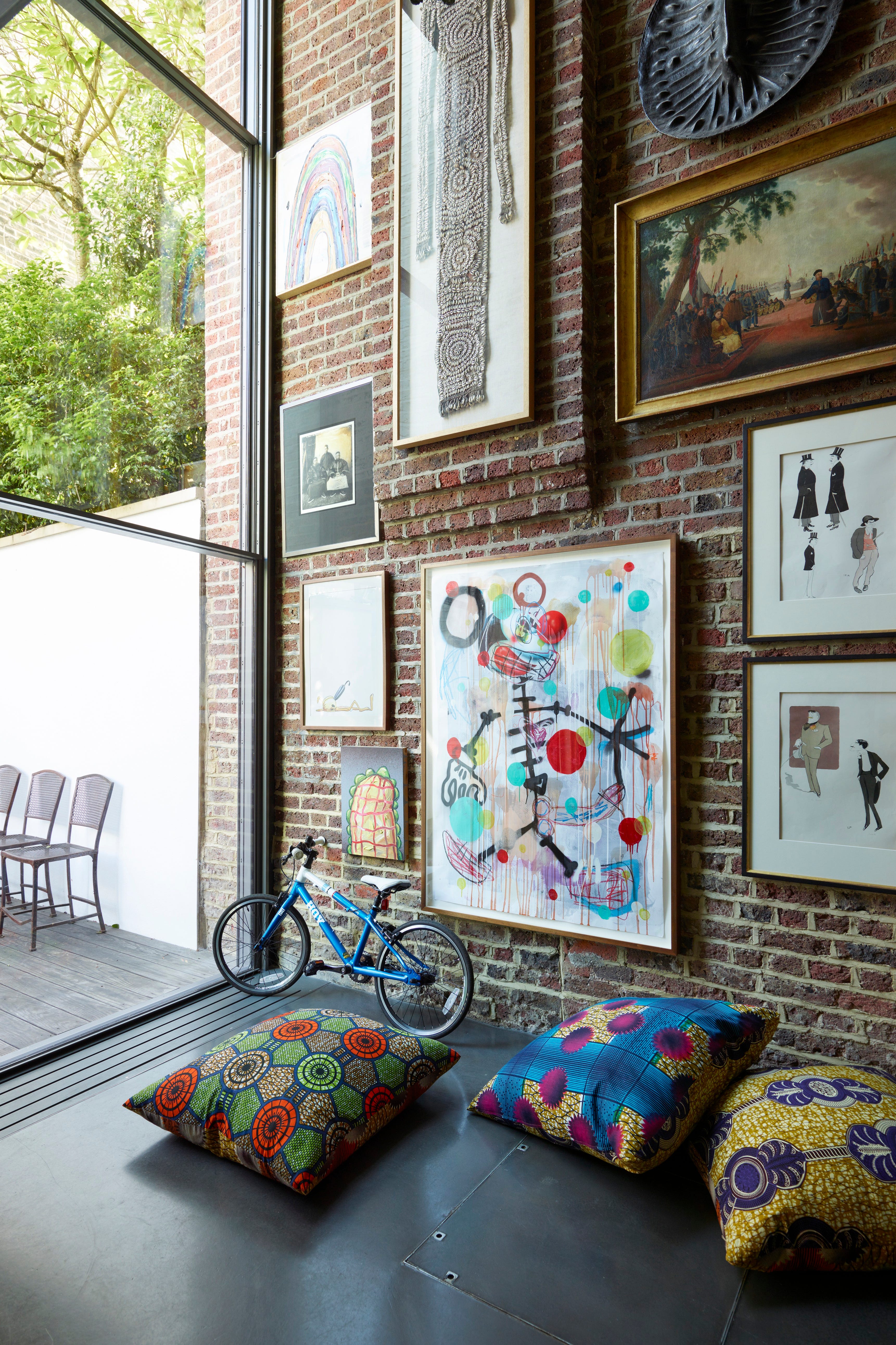 Curated gallery wall on exposed brick — mixed custom art pieces of different scales, plus vibrant patterned floor pillows — the art-in-situ approach to guest-space personality.