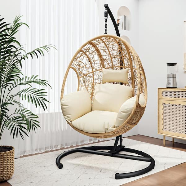 Wicker egg chair with cream cushions suspended from a black steel stand in a bright living room with palm plants and a rattan sideboard — the indoor hanging-furniture trend in situ.