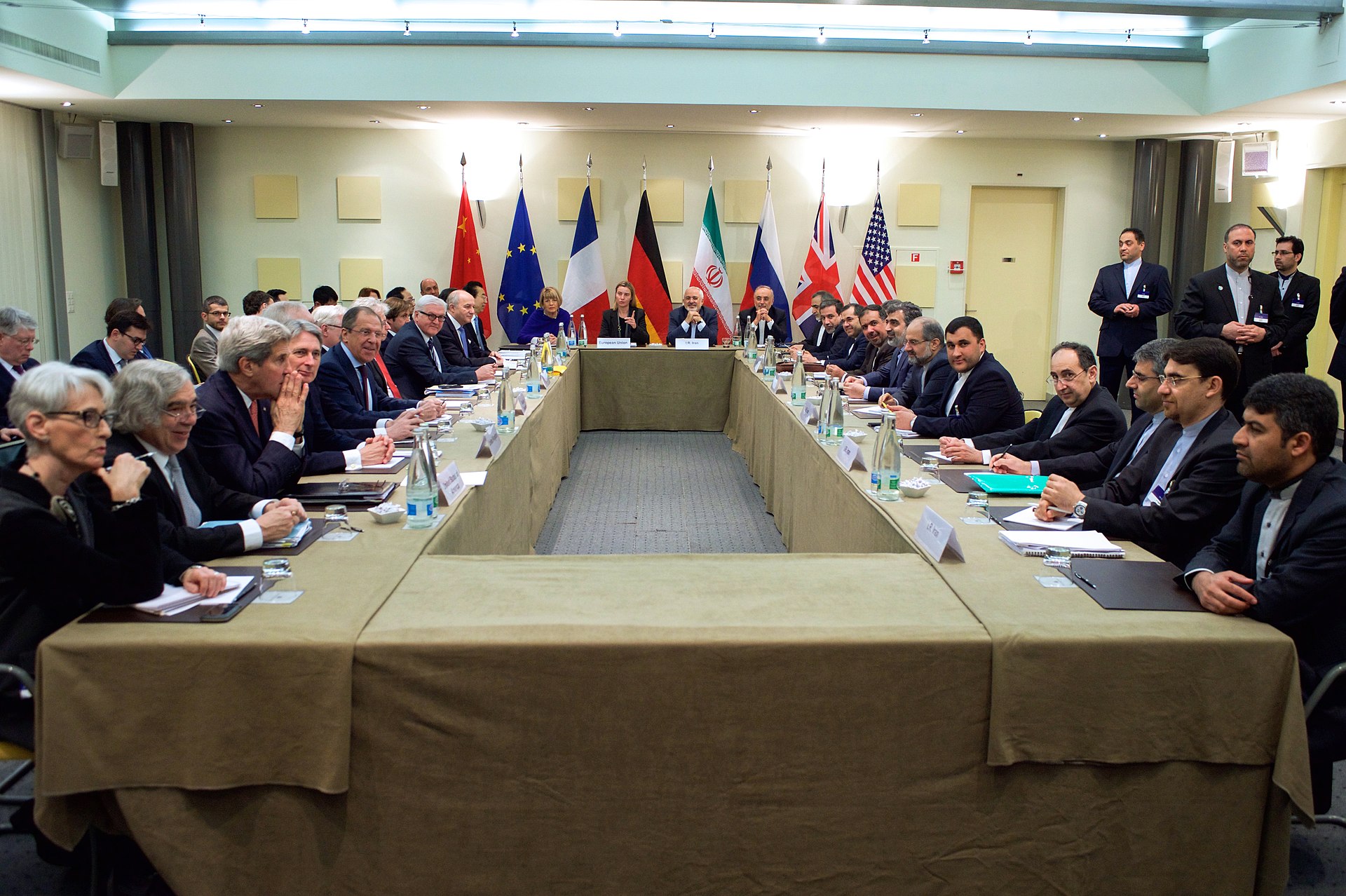 Multilateral in-person negotiation — diplomats from the US, EU, UK, France, Germany, Russia, China, and Iran at a formal negotiating table with national flags behind them.